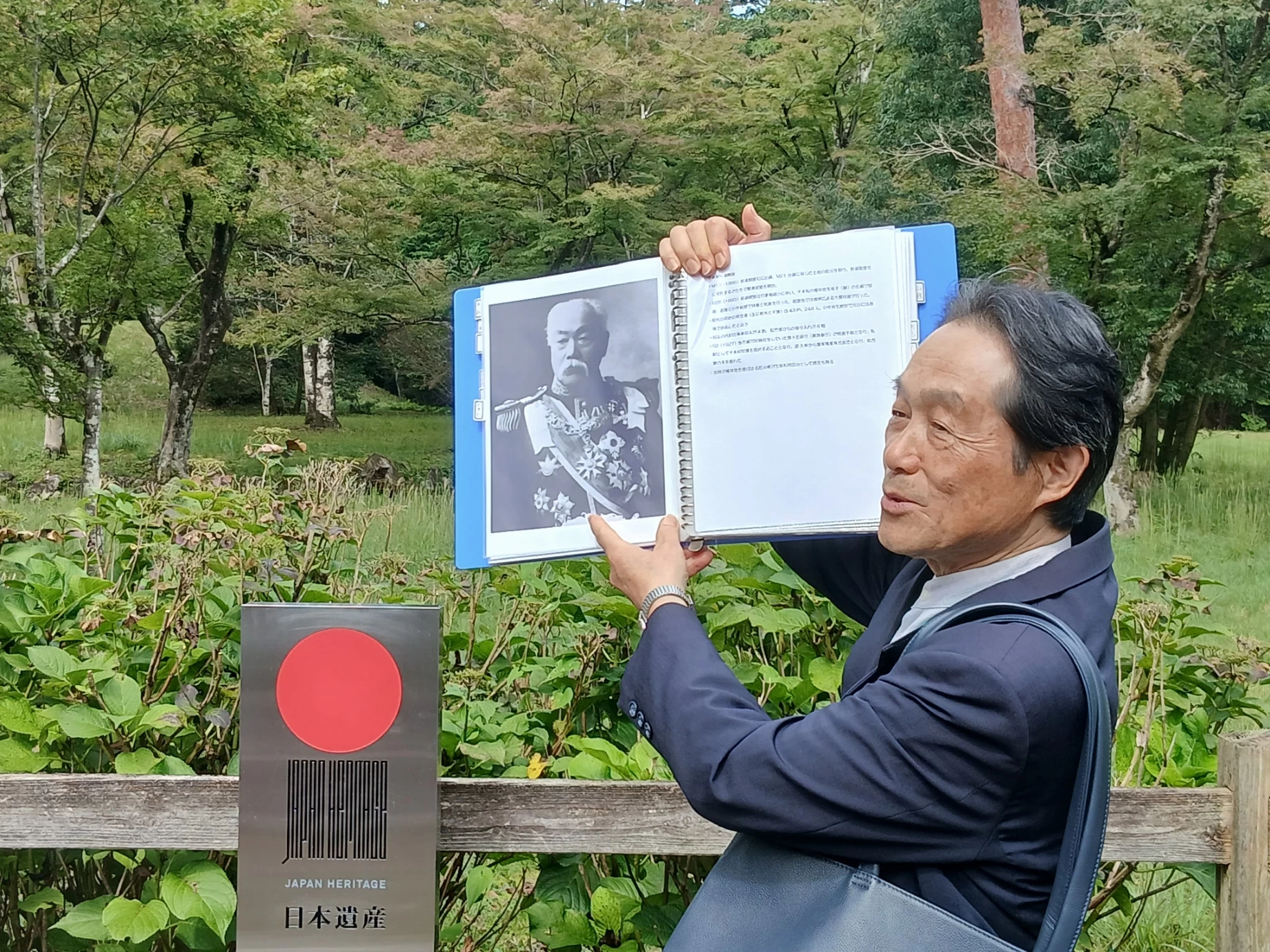 日本遺産の標柱の前で松方正義の肖像を見せるガイド